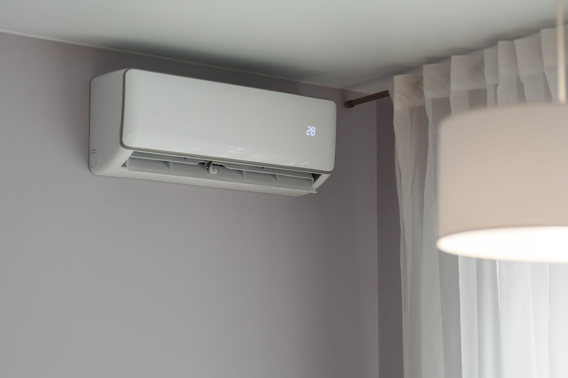 Climatiseur mural blanc à côté d'une fenêtre avec des voilages et un lampadaire dans une pièce grise.