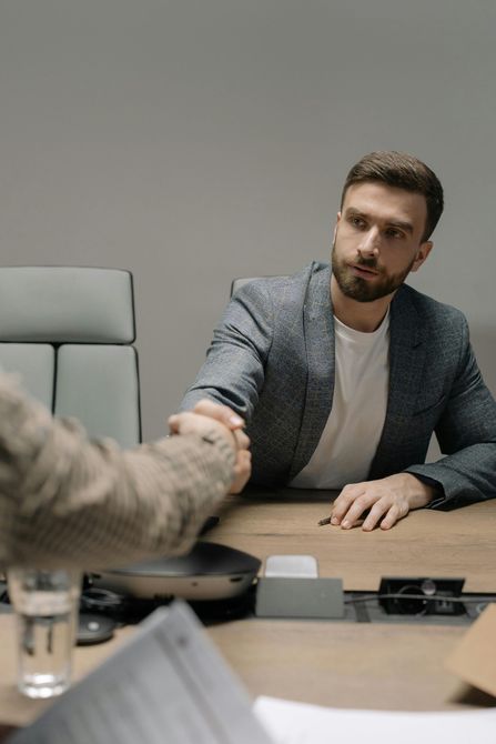 Hombre con blazer estrechando manos desde un escritorio. Ambiente de oficina, expresión seria.