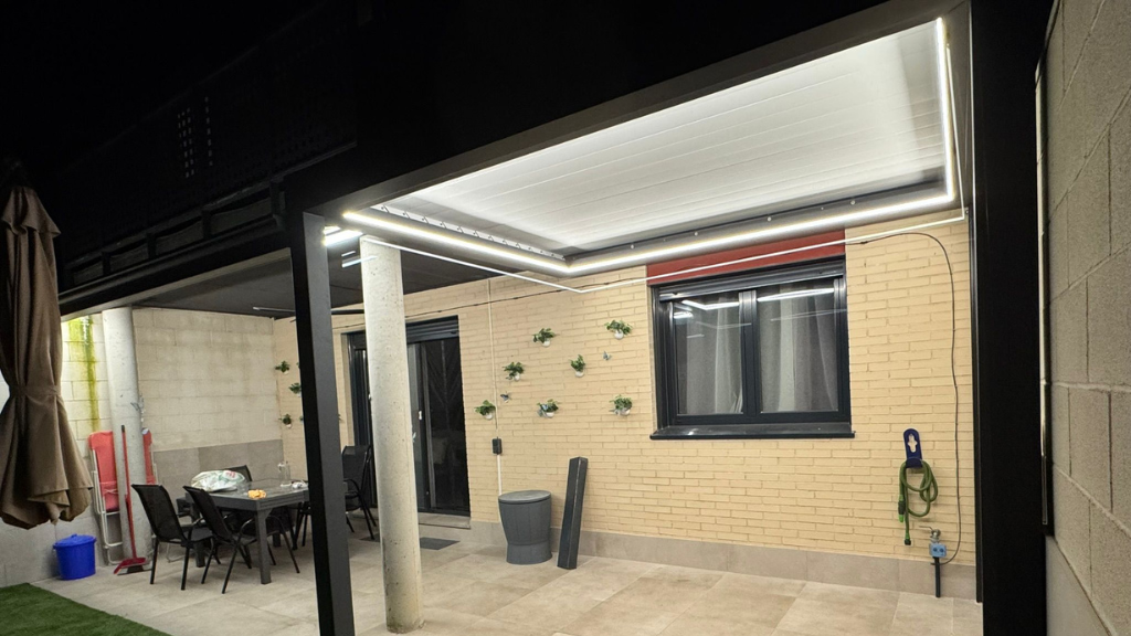 Patio iluminado con pared de ladrillo, ventana, papeleras, plantas colgantes y zona de estar cubierta bajo luces brillantes.