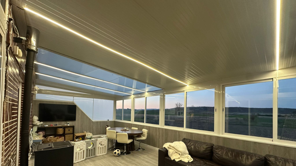 Salón moderno con ventanas panorámicas, asientos acolchados y luces cálidas en el techo con vistas a un paisaje al atardecer.
