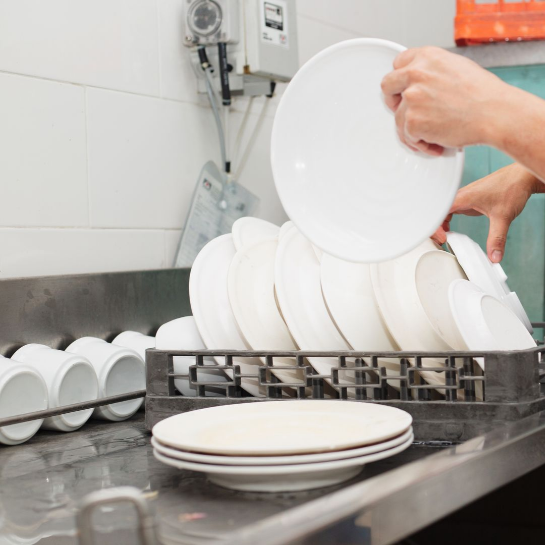 Des mains empilent des assiettes blanches dans un égouttoir à vaisselle près de l'évier d'une cuisine professionnelle.
