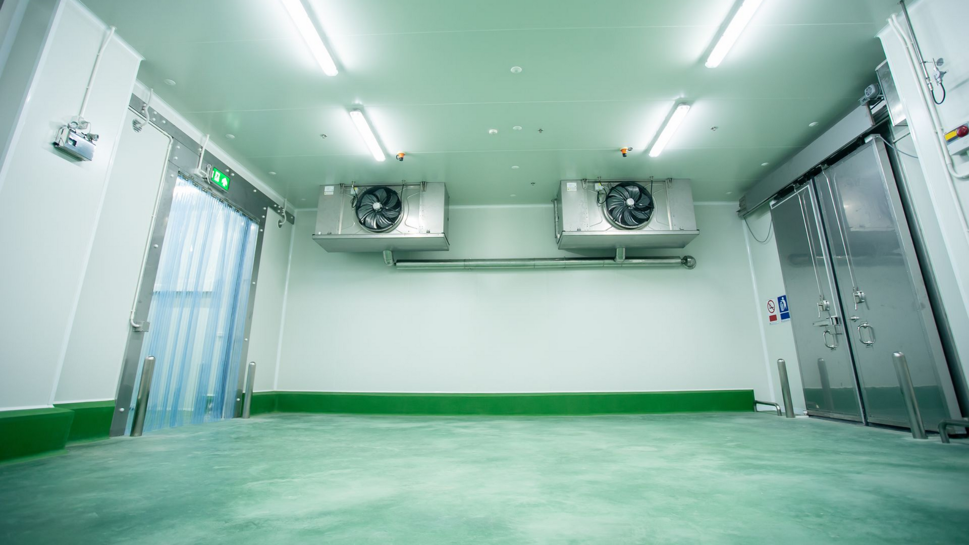 Chambre froide vide, blanche, avec sol vert, ventilateurs muraux et porte vitrée.
