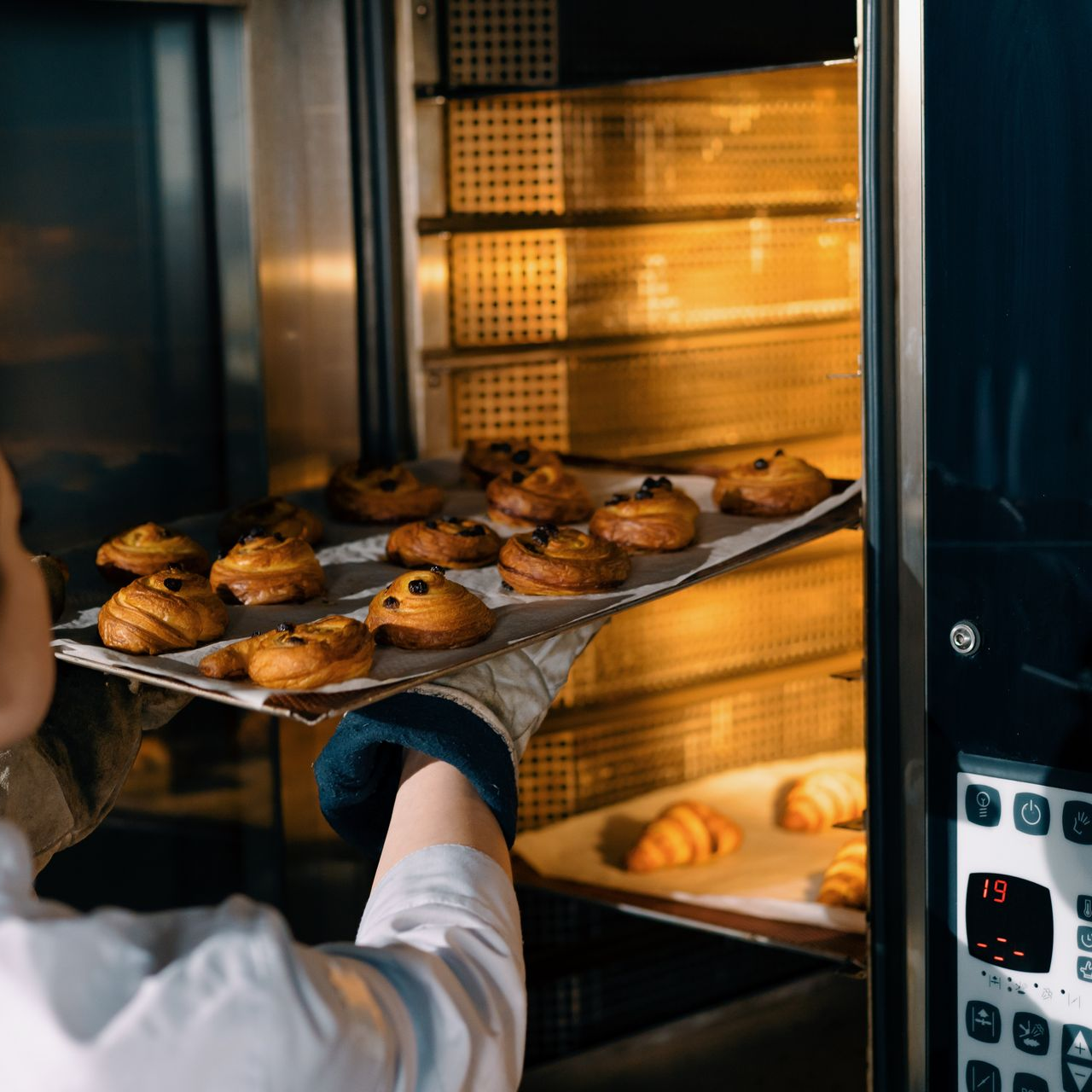 Un boulanger glisse une plaque de muffins dans un four, dont la lumière chaude et le panneau de commande sont visibles.