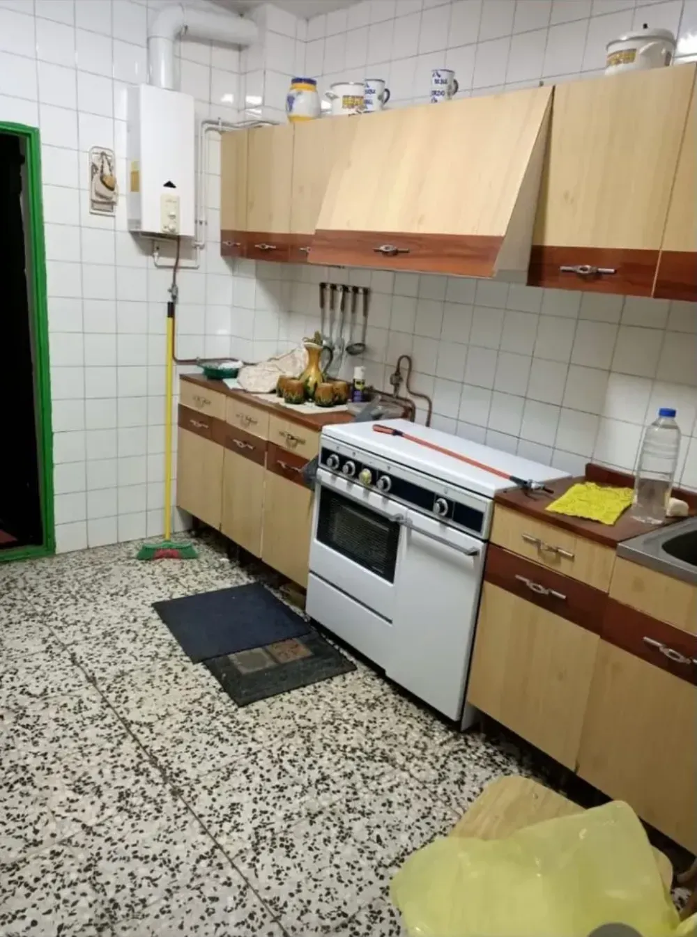 Cocina pequeña con armarios de madera clara, azulejos blancos, una estufa y un suelo de baldosas moteadas.