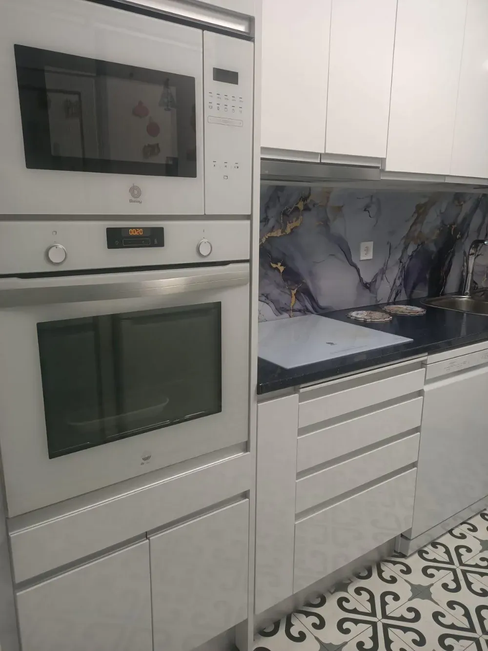 Cocina moderna blanca con horno y microondas empotrados, armarios blancos y salpicadero de mármol gris.