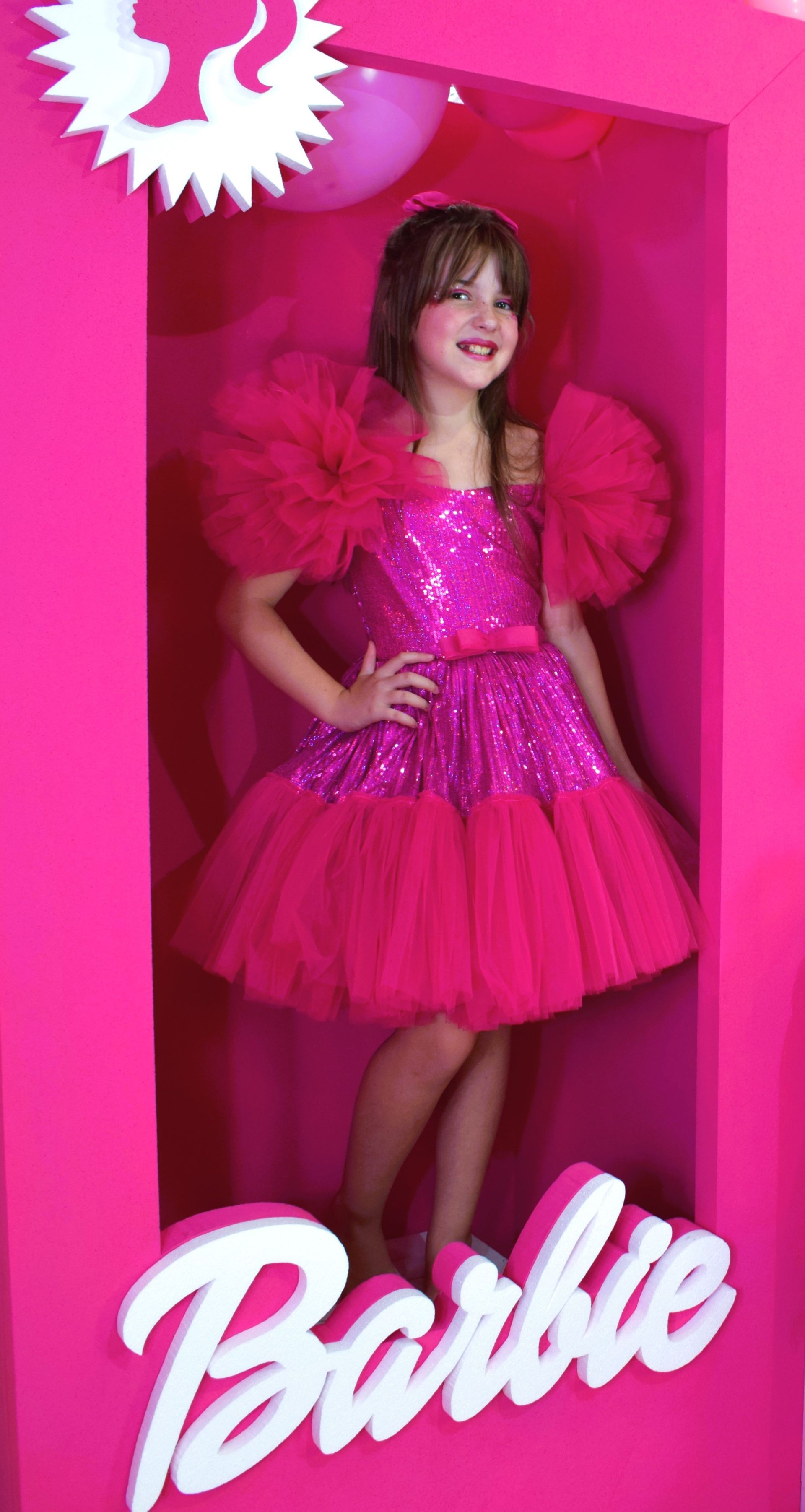 Una niña con un vestido rosa está parada dentro de una caja de muñecas Barbie rosa.