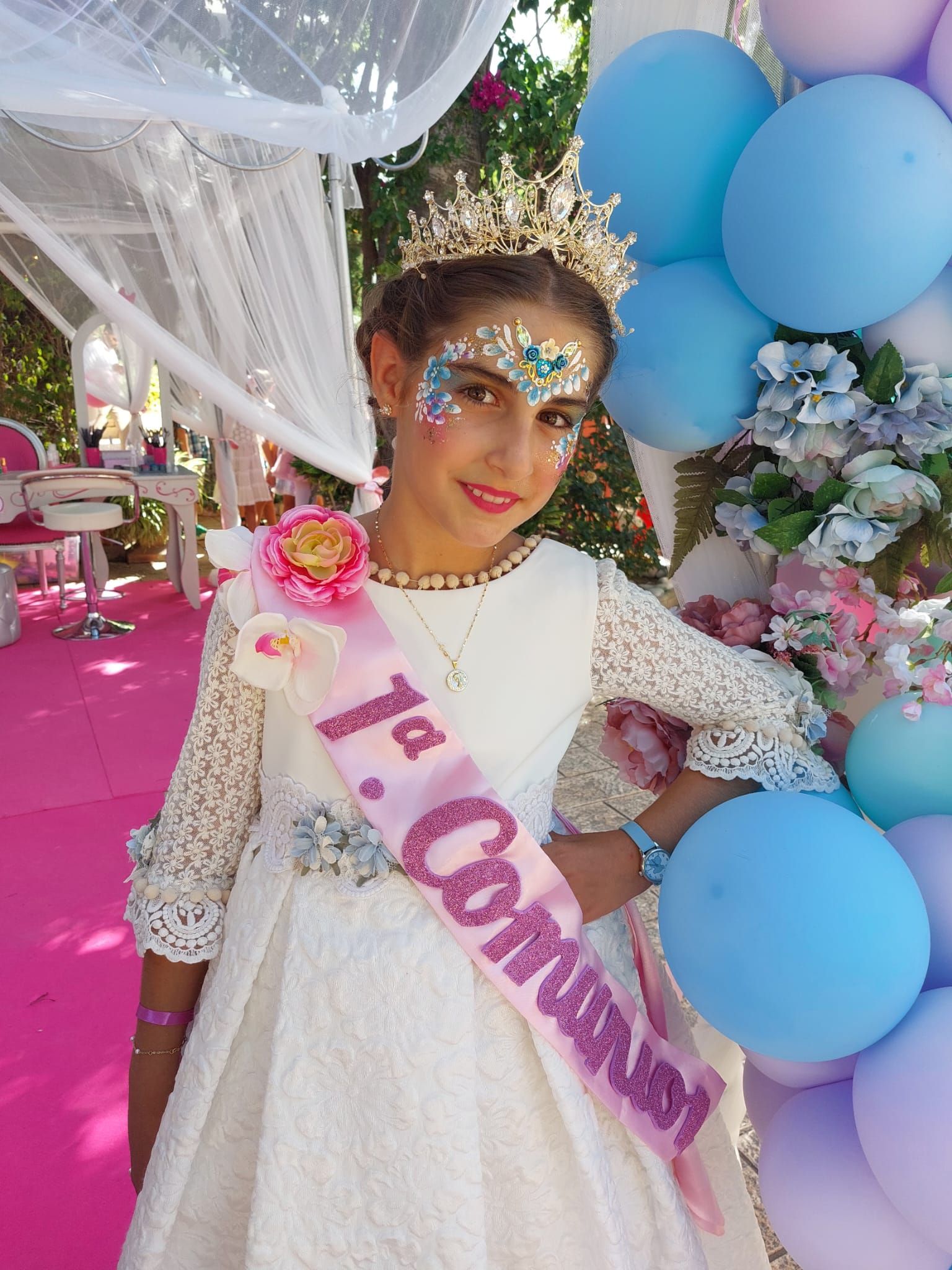 Una niña con un vestido blanco y una corona está parada frente a globos.