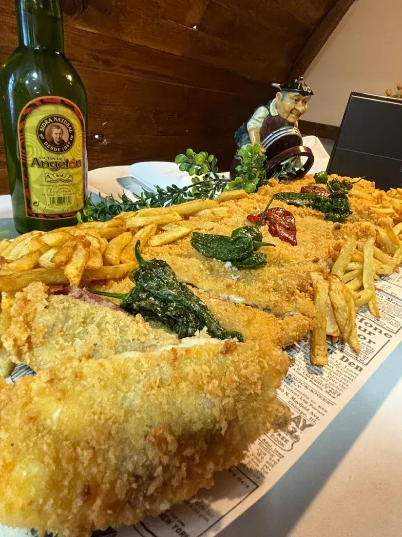 Gran plato de pescado frito con patatas fritas y verduras, servido en una bandeja con una botella de cerveza al fondo.