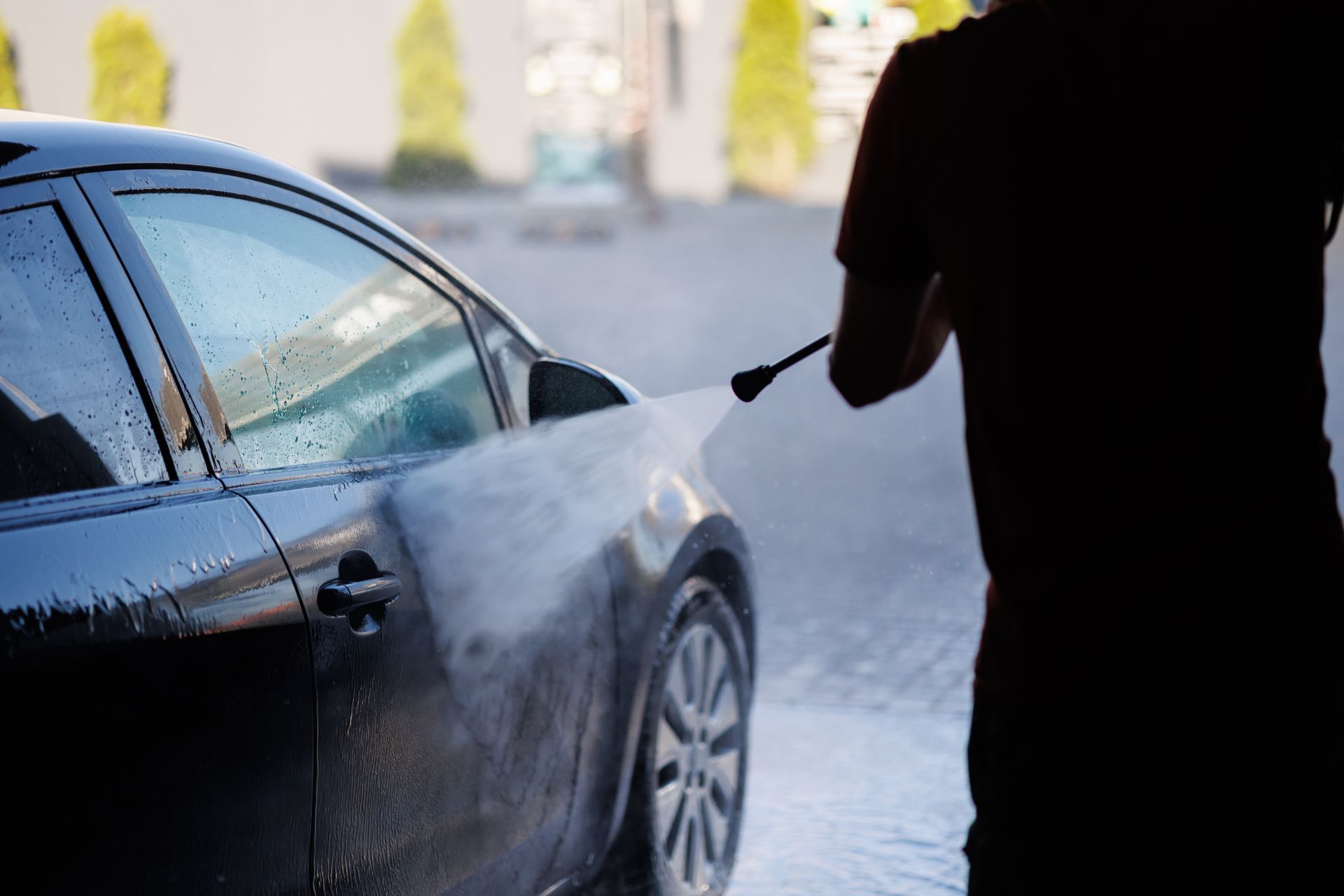 Une personne lave une voiture noire avec un nettoyeur à haute pression à l'extérieur.