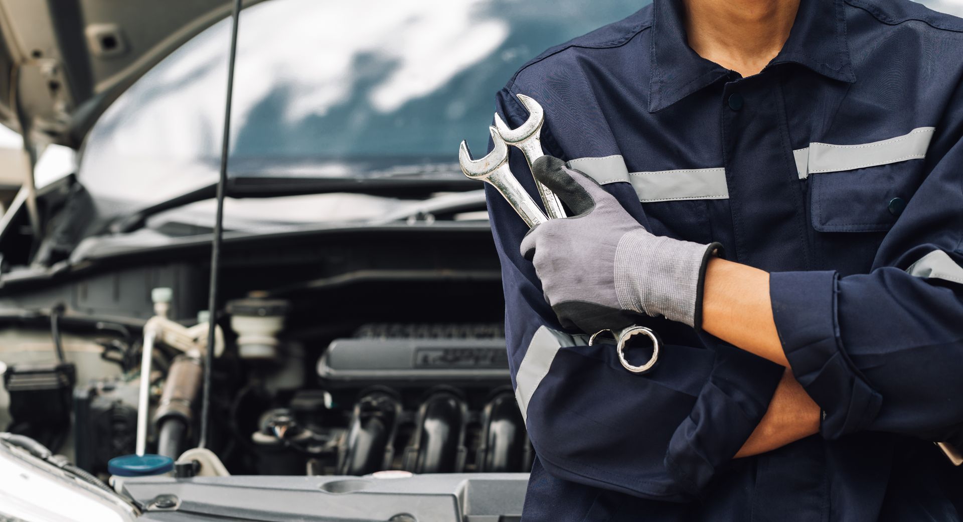 Mécanicien en uniforme bleu, tenant des clés devant une voiture avec un capot ouvert.