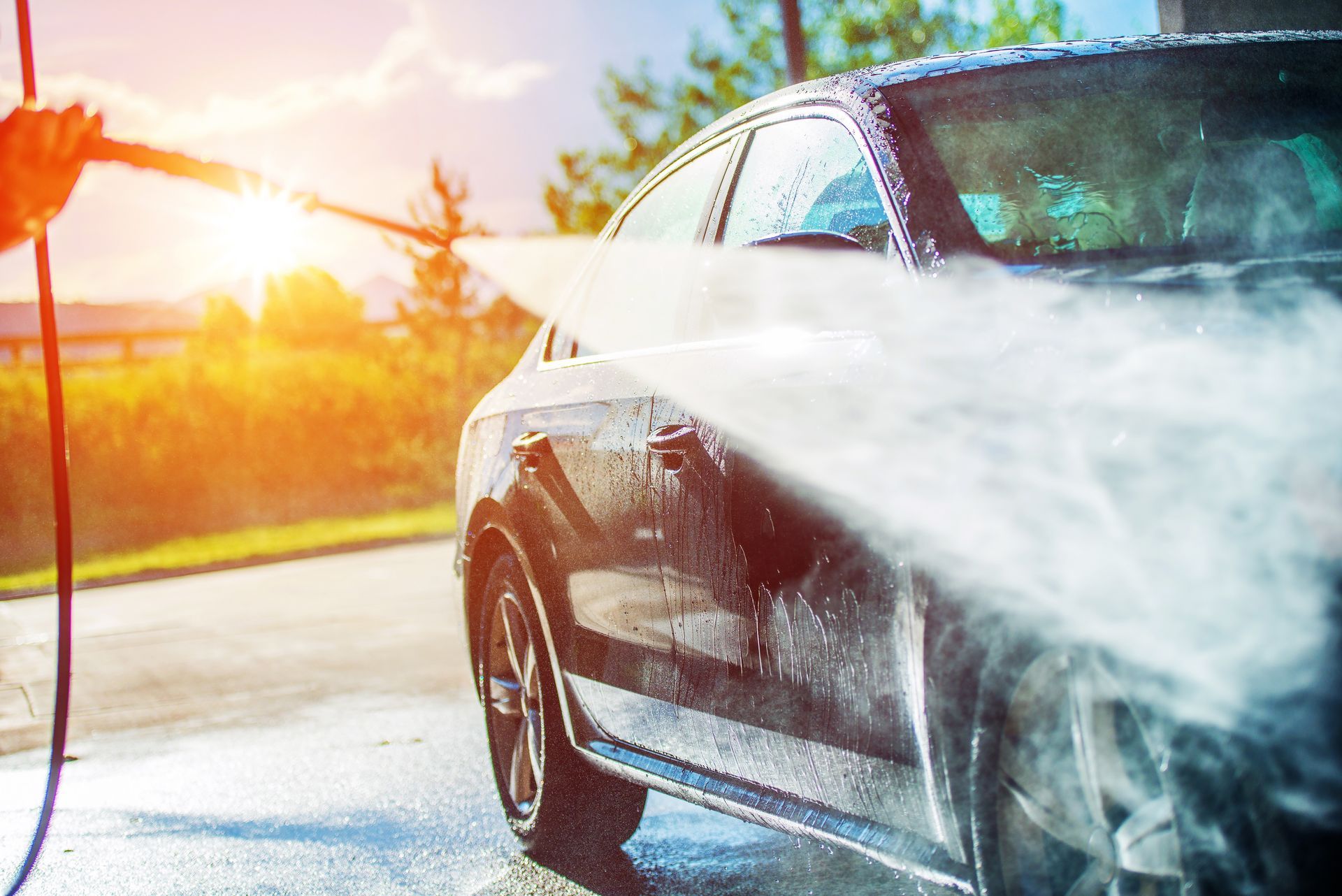 Une personne lave une voiture sombre avec un nettoyeur à haute pression en plein soleil.