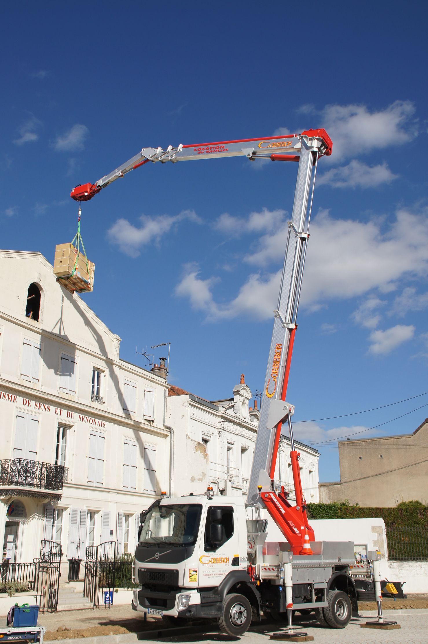 Grue montée sur camion soulevant un bloc de pierre de la façade d'un bâtiment sur fond de ciel bleu éclatant.