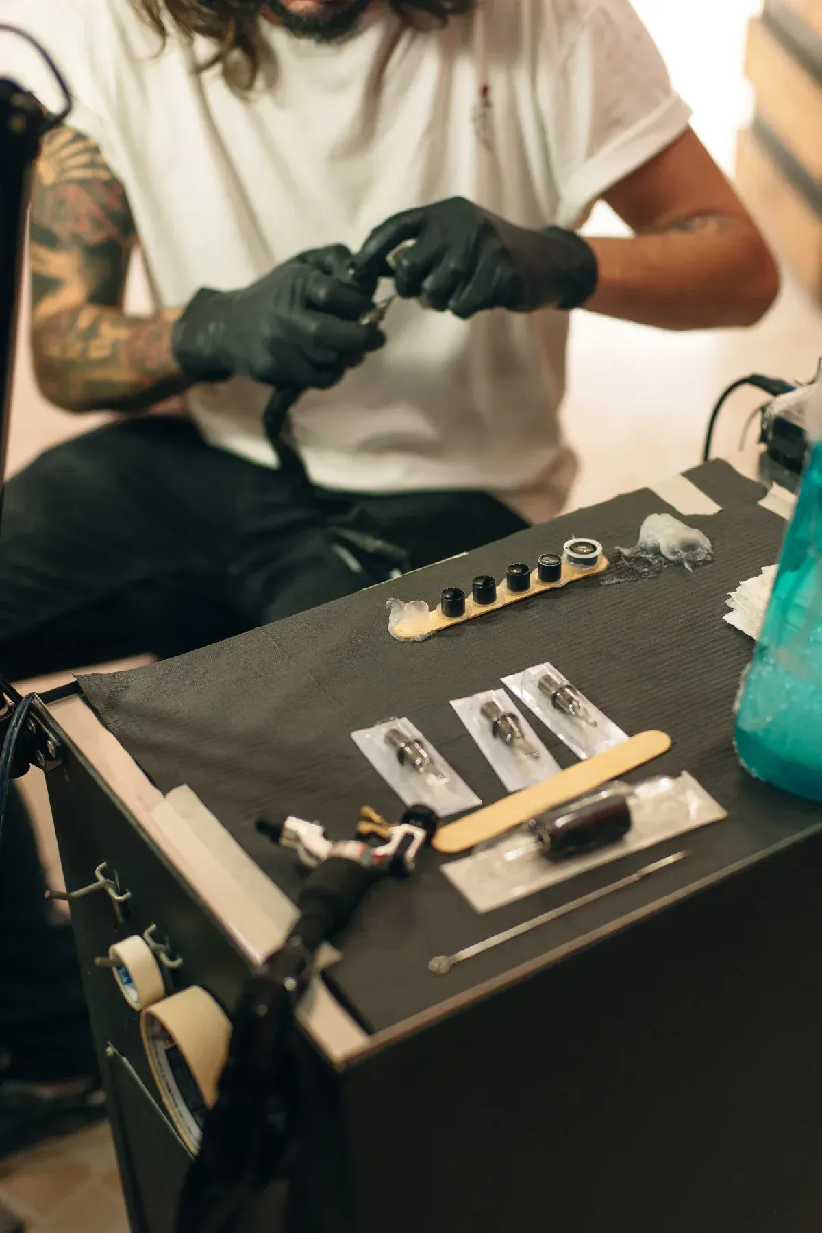 Un artista del tatuaje que viste guantes negros prepara el equipo en una mesa con agujas, tinta y una máquina de tatuar.