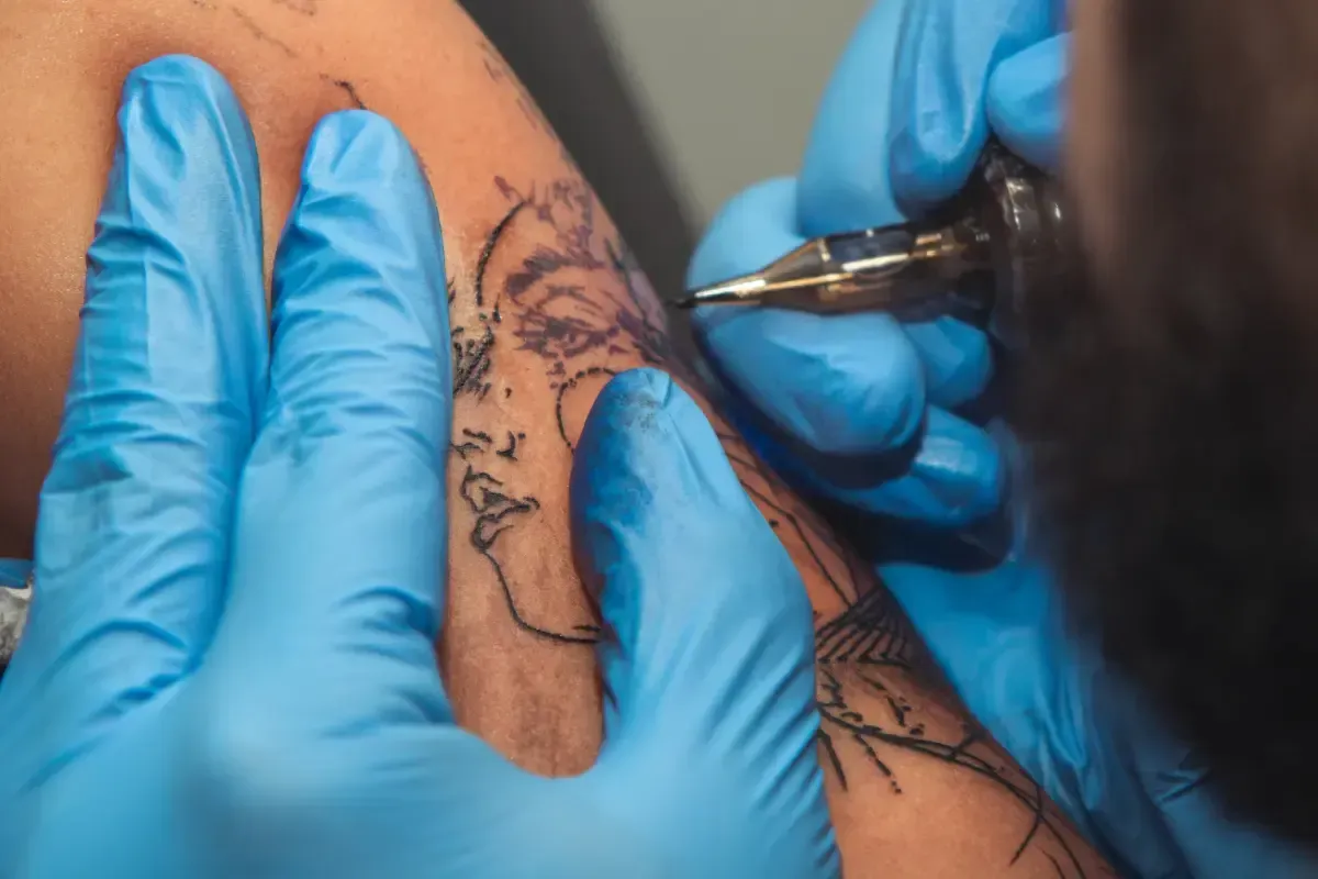 Manos con guantes azules tatuando el rostro de una mujer en un brazo.
