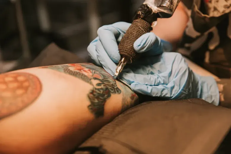 El brazo de una persona se está tatuando; la mano enguantada del artista sostiene una pistola de tatuajes