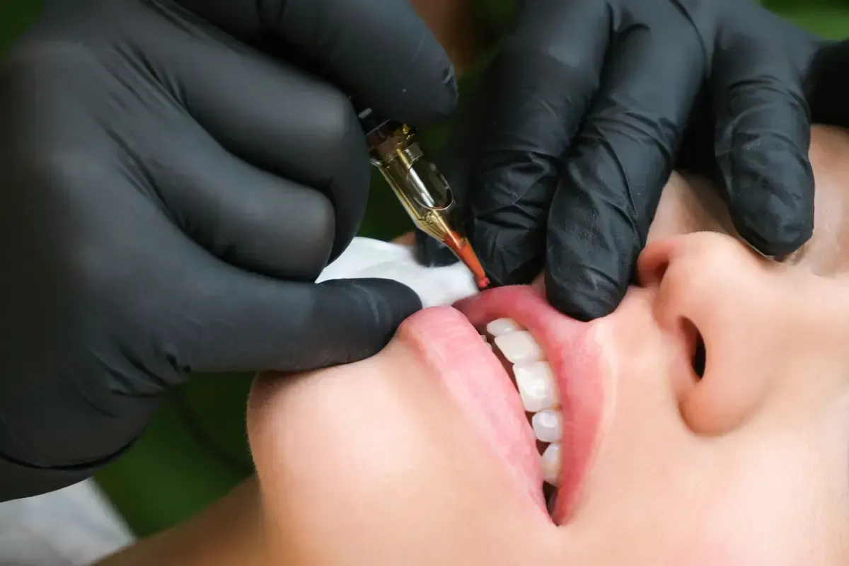 Persona recibiendo un tatuaje de labios, manos del artista con guantes negros utilizando una máquina de tatuar en los labios.