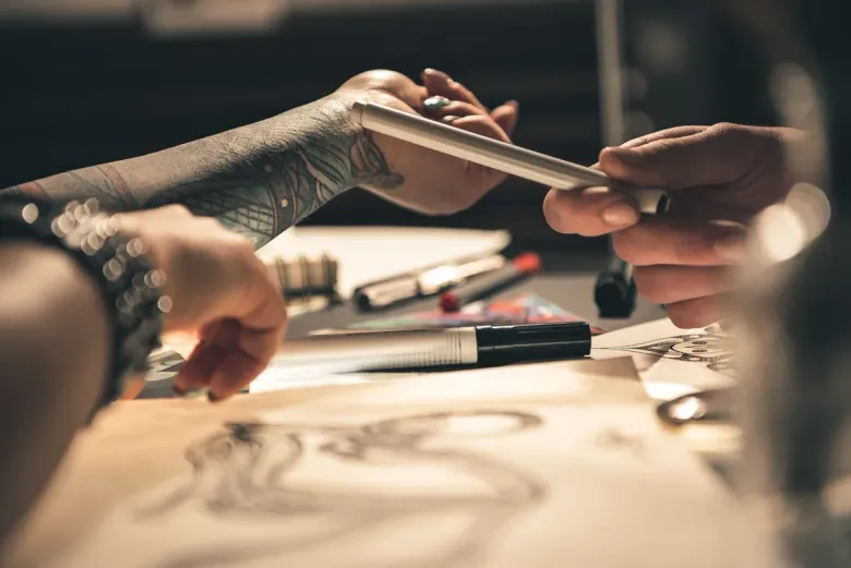 Persona con tatuaje en el brazo recibe un bolígrafo para dibujar. Estudio con bolígrafos y dibujos.