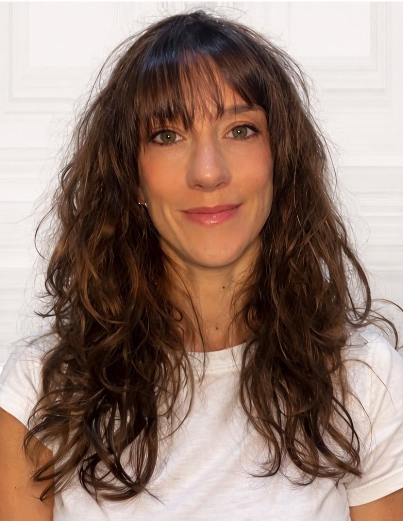 Mujer de cabello oscuro mirando a la cámara con una leve sonrisa. Al aire libre, con ramas de árboles al fondo.
