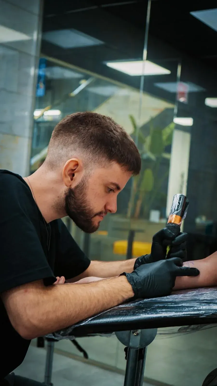 Artista del tatuaje con pelo corto y barba, con guantes, tatuando el brazo de un cliente en un estudio de tatuajes.