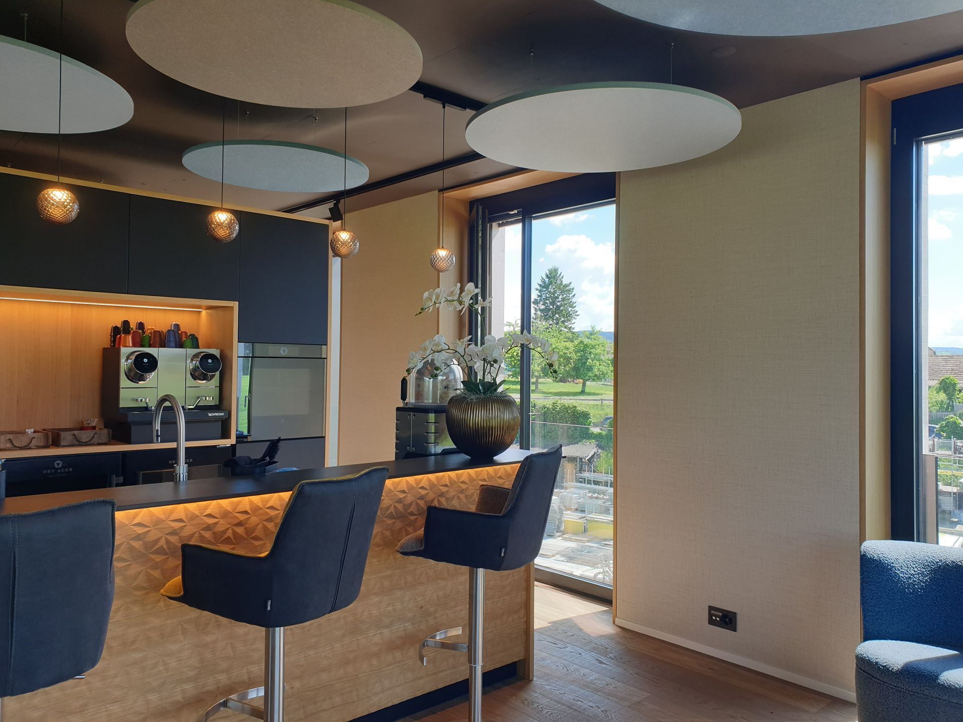 Moderne Küche mit Barhockern, dunkler Arbeitsplatte und Blick durch ein Fenster auf eine grüne Landschaft.