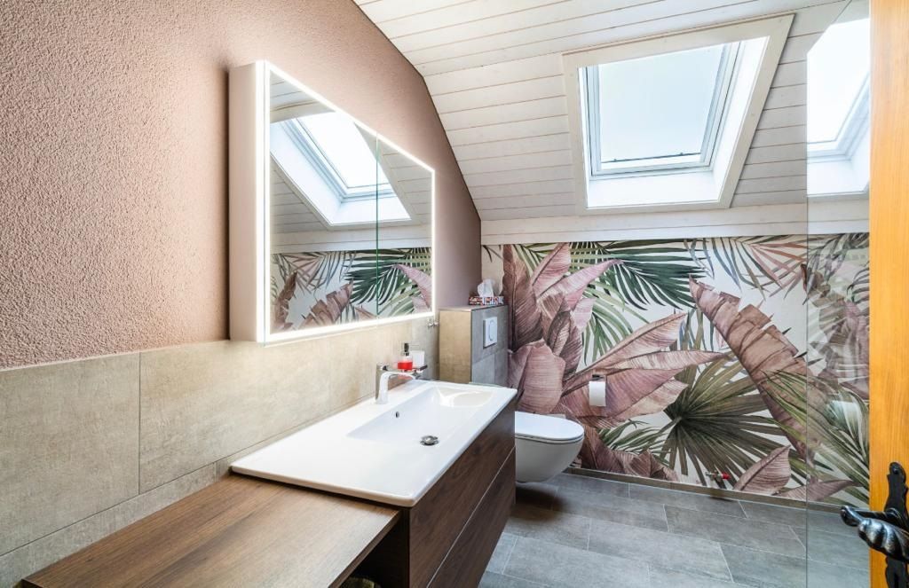 Badezimmer mit rosa Wänden, tropischer Tapete, Waschbecken, Toilette und Oberlichtern.