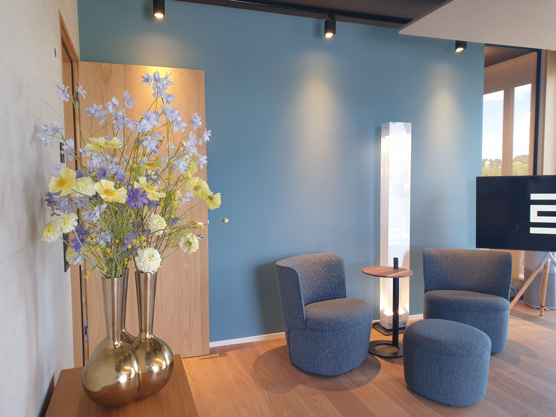 Zimmer mit blauer Wand, Stühlen, Stehlampe, Blumen in goldener Vase, Holztür und Tisch.