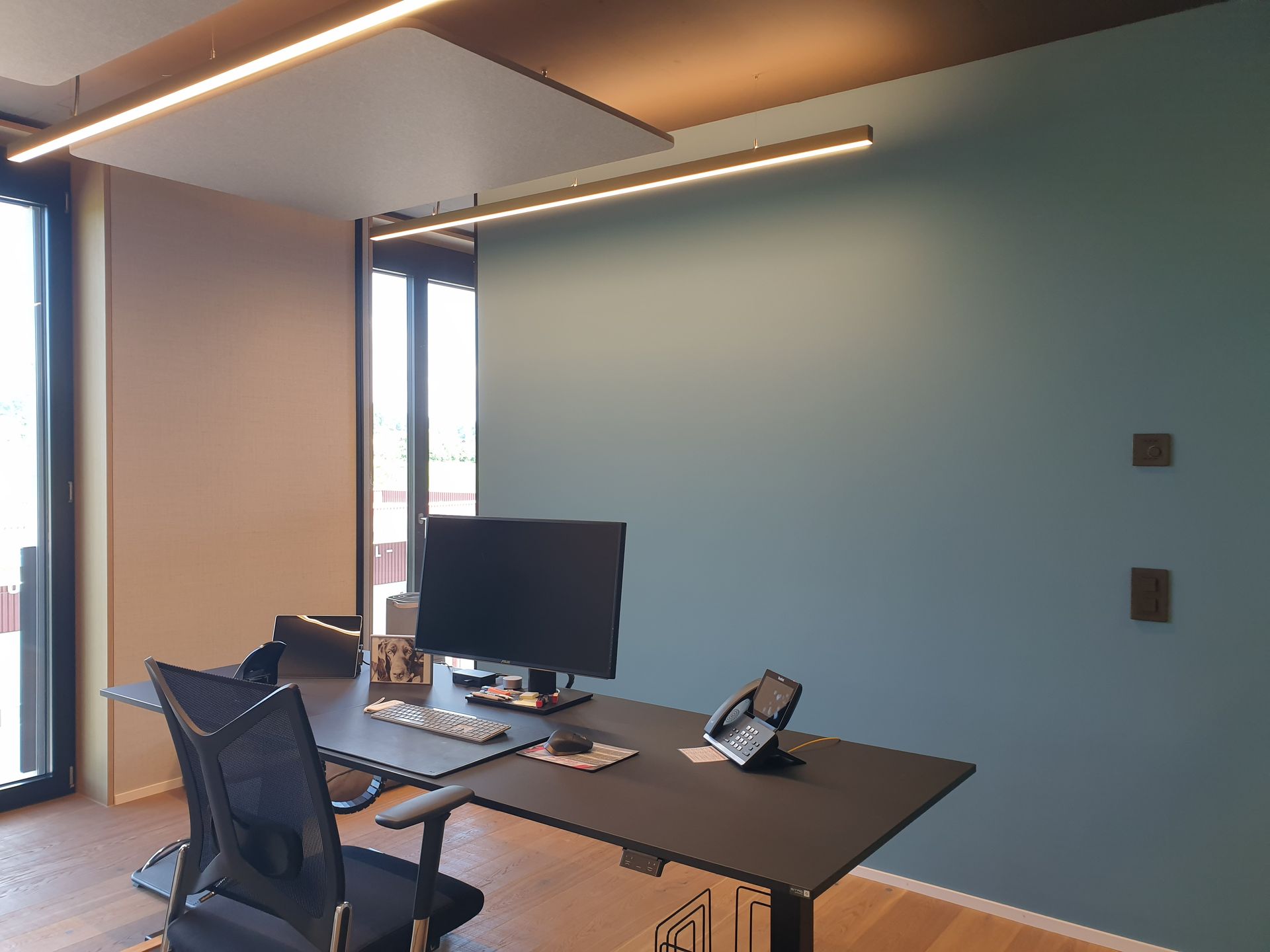 Büro mit blauer Akzentwand, Schreibtisch mit Computer, schwarzem Stuhl und moderner Beleuchtung.