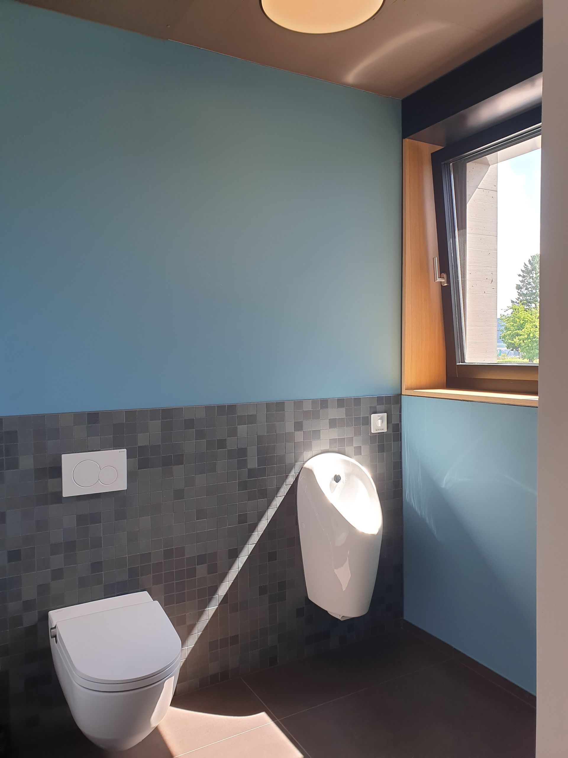 Blau-graues Badezimmer mit Toilette und Urinal neben einem Fenster.