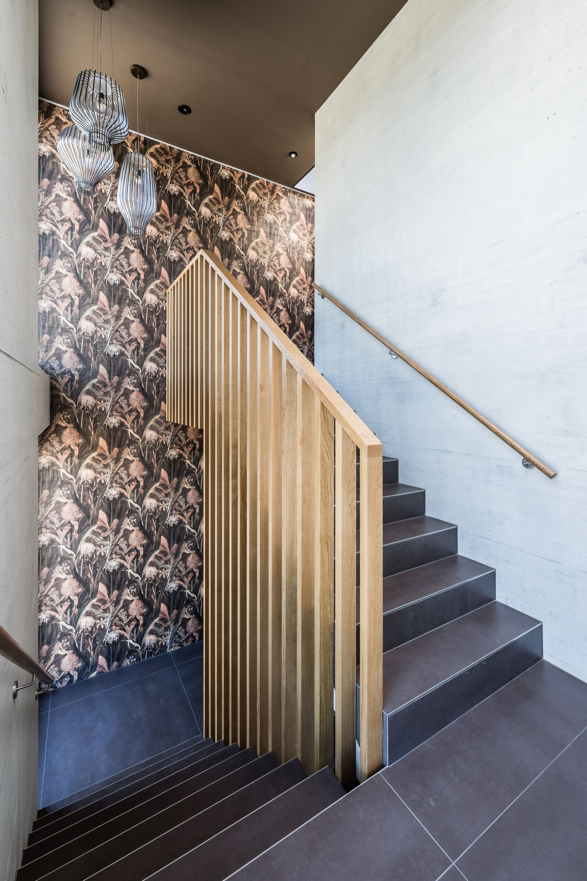 Treppe mit Holzgeländer und dunkel gefliesten Stufen vor gemusterter Tapete.