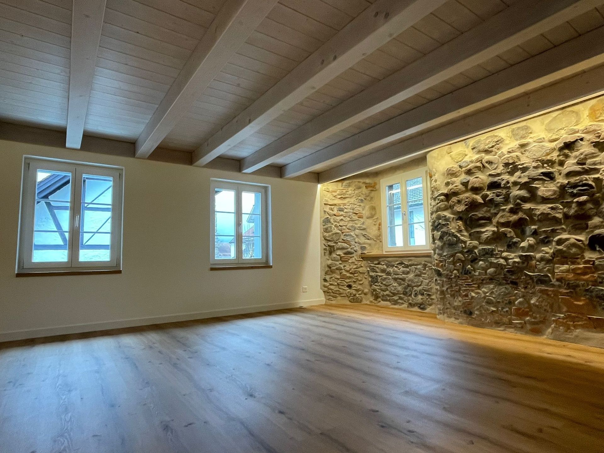 Leerer Raum mit Holzboden, freiliegender Balkendecke, Steinmauer und Fenstern.
