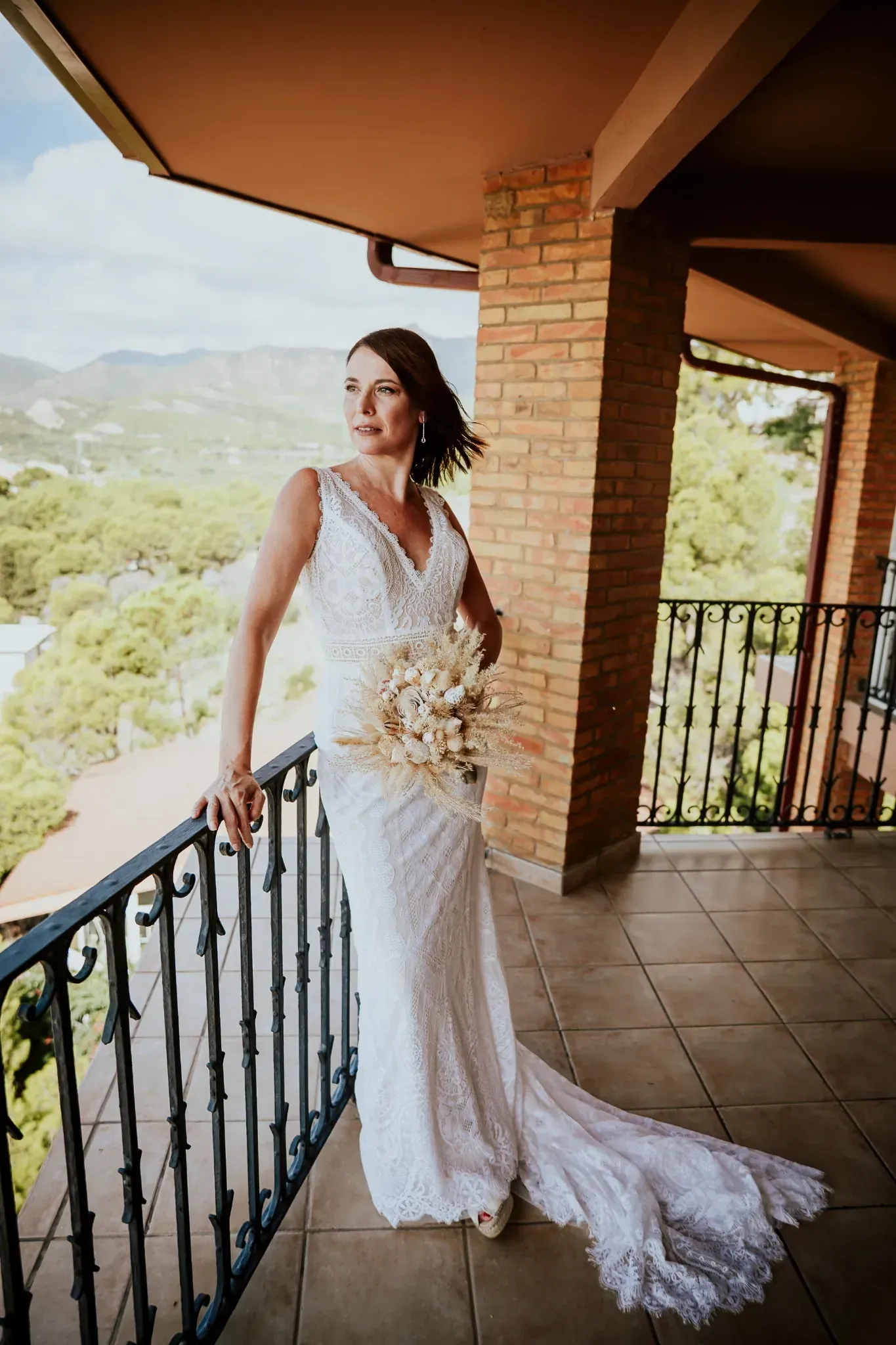 Mujer con vestido de novia de encaje blanco en el balcón, sosteniendo flores, con vistas a un paisaje.