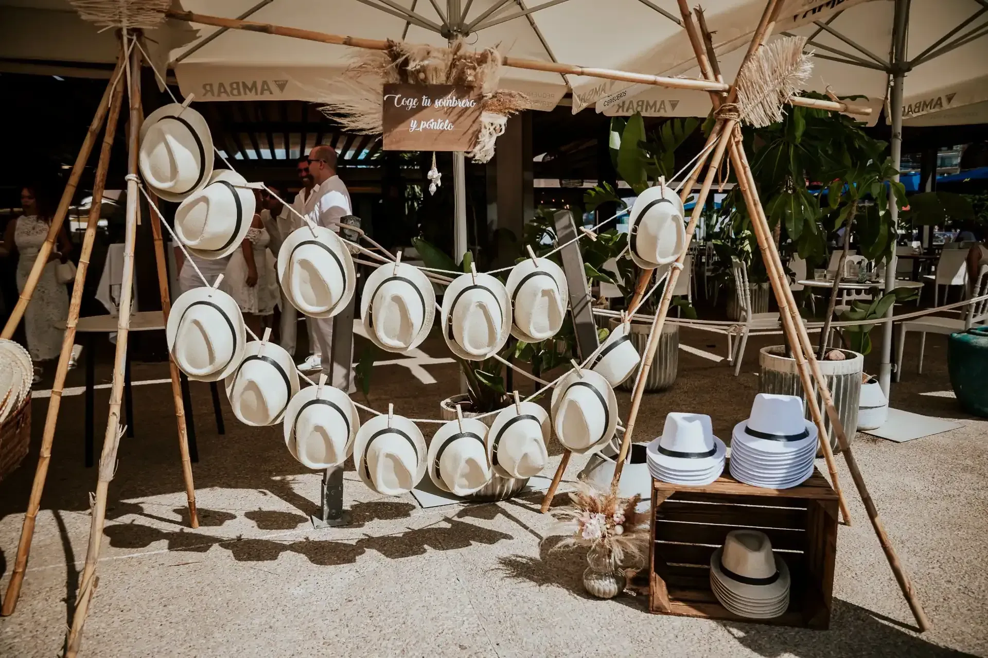 Exhibición de sombreros en un evento al aire libre con un marco de madera y un cartel, rodeado de sombrillas y plantas.