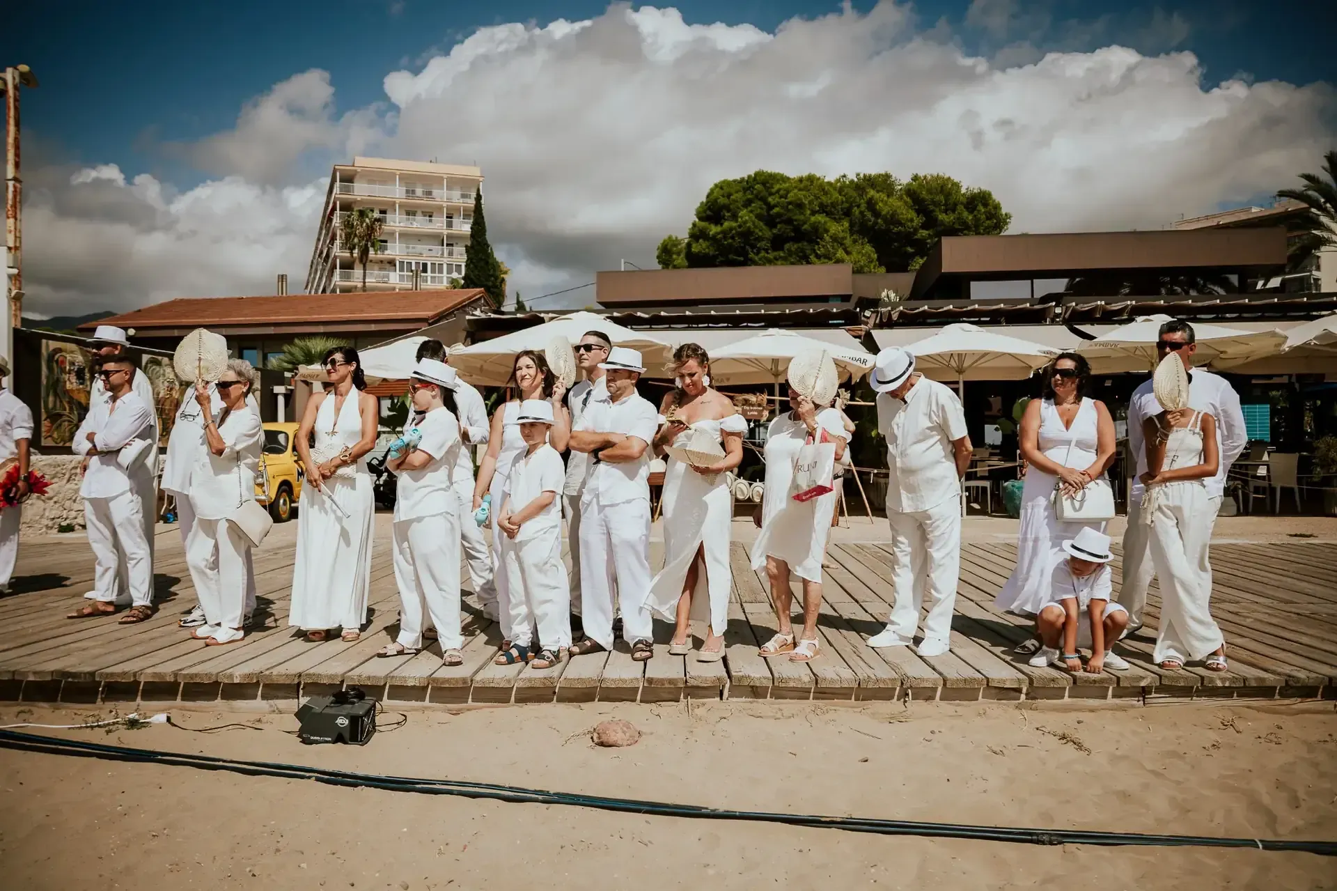 Personas vestidas de blanco de pie en un paseo marítimo cerca de una playa con edificios y nubes en el fondo.