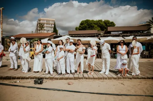 Personas vestidas de blanco de pie en un paseo marítimo cerca de una playa con edificios y nubes en el fondo.