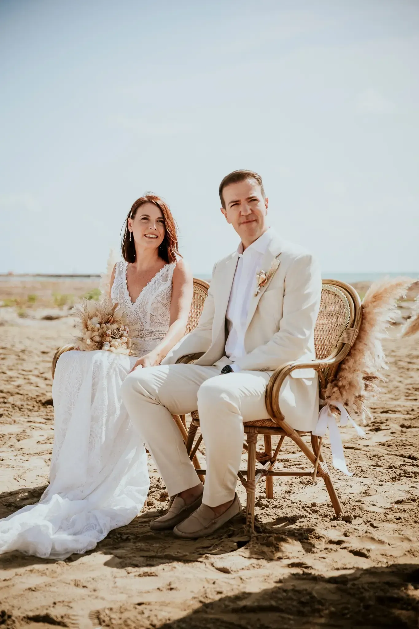 Una pareja con traje de boda sentada en una silla de mimbre en la playa. Cielo soleado y despejado.
