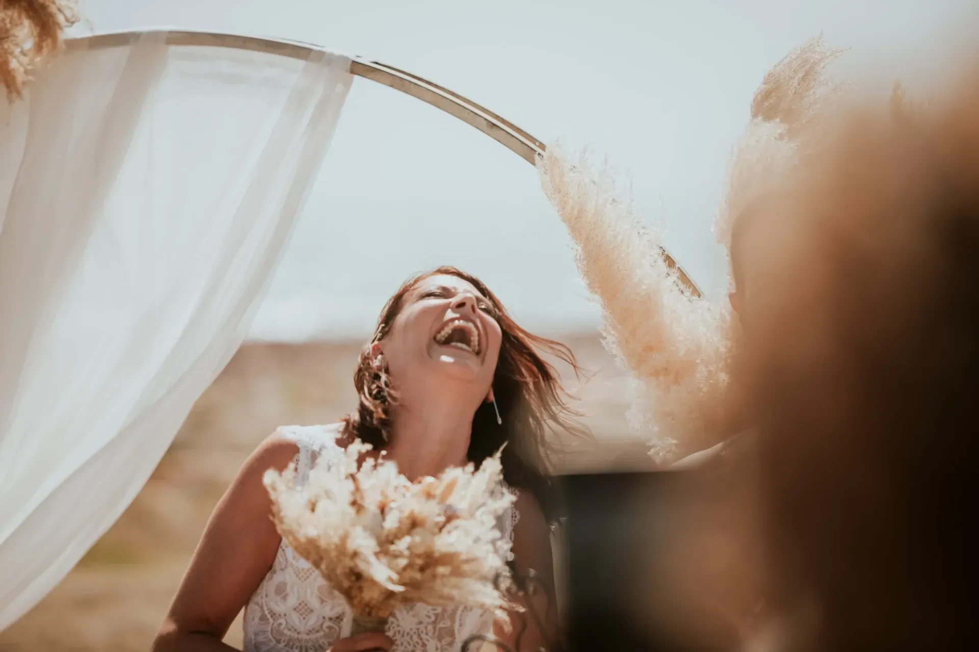 La novia se ríe alegremente, sosteniendo el ramo bajo un arco cubierto al aire libre.
