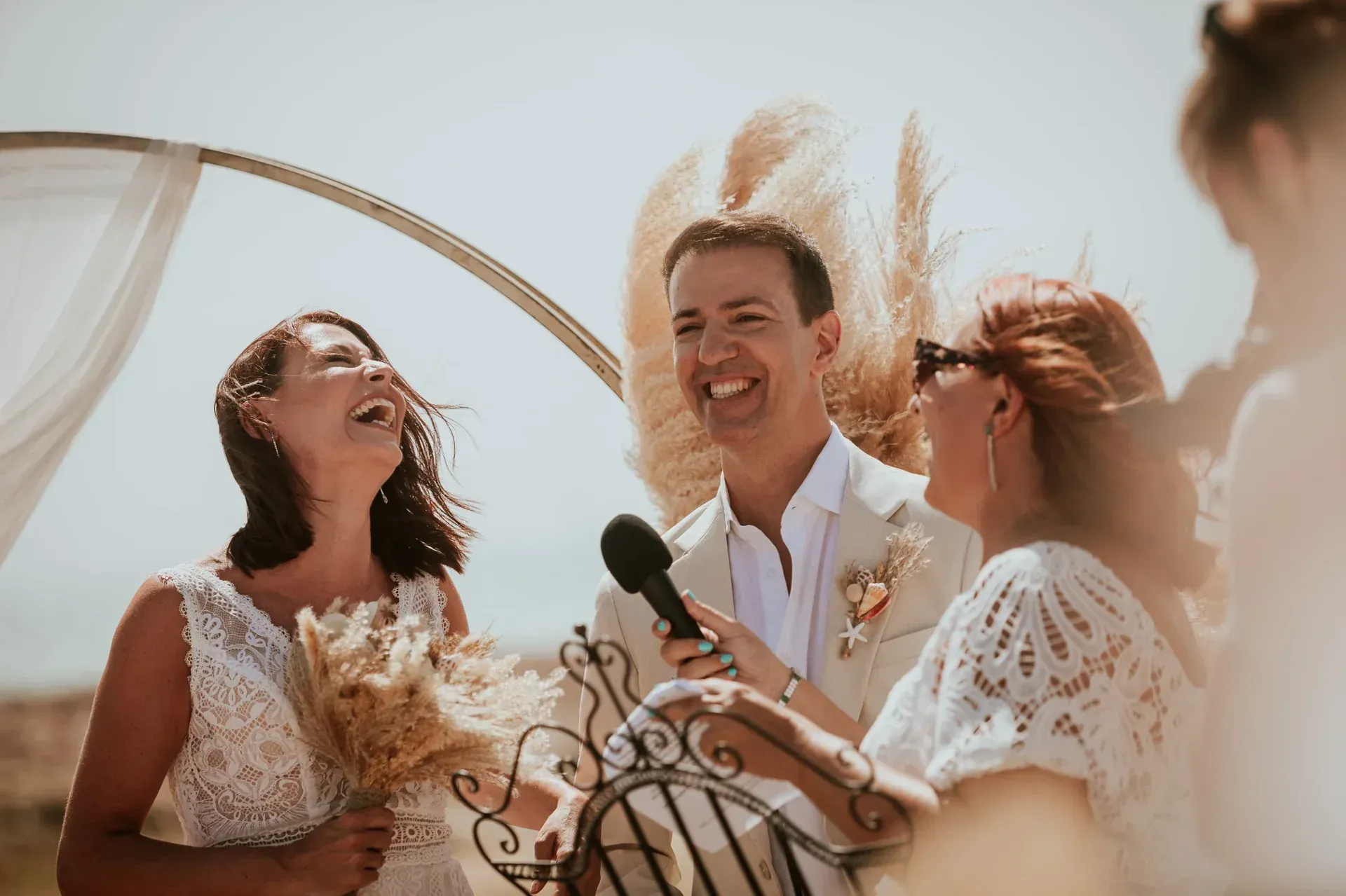 La novia, el novio y el oficiante se ríen durante una ceremonia de boda al aire libre; tonos neutros, entorno soleado.