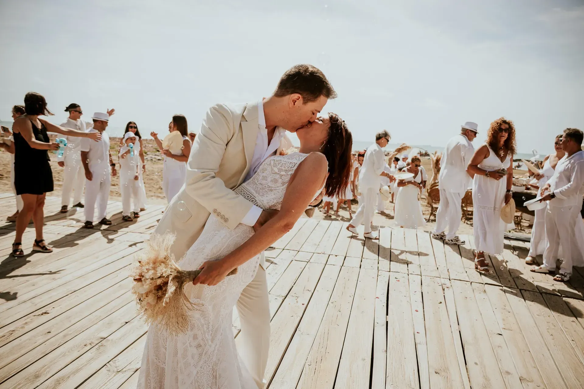 Pareja besándose en una pasarela de madera, rodeada de gente vestida de blanco, en un entorno soleado al aire libre.