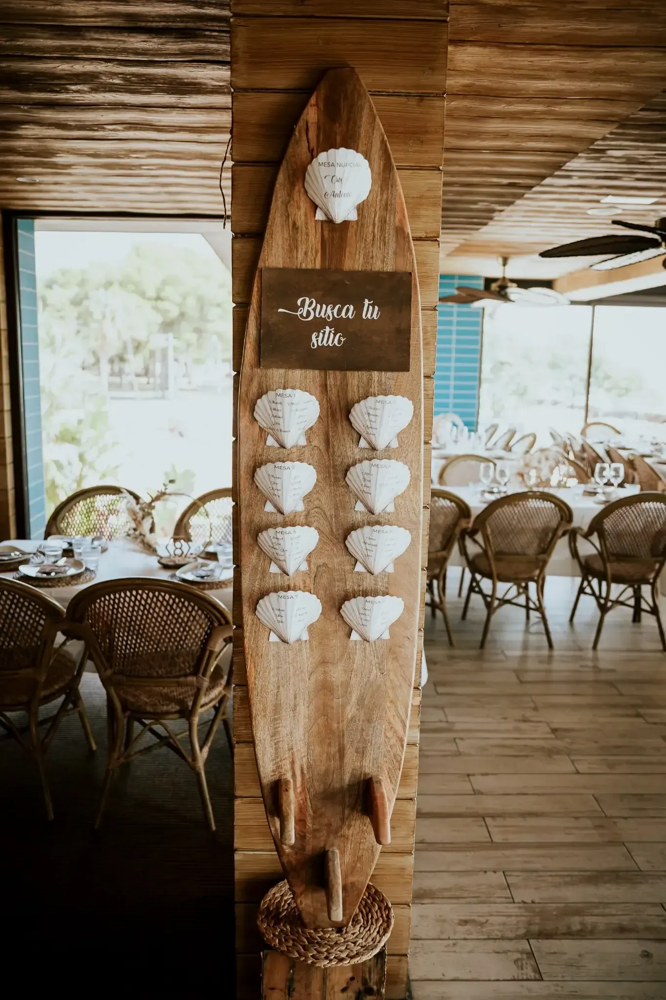 Tabla de surf de madera con tarjetas de asientos para invitados, colocadas cerca de mesas y sillas en un lugar.