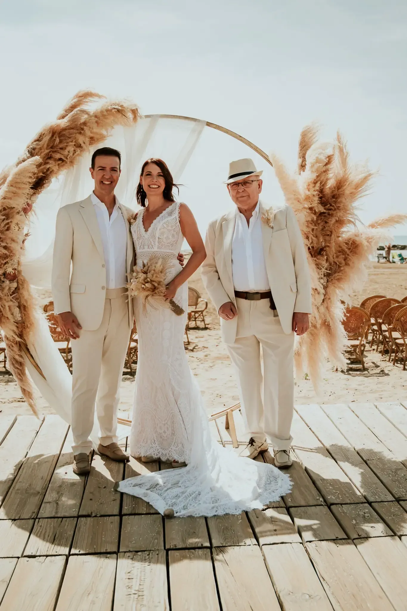 La novia, el novio y el hombre, con trajes beige, posan en una ceremonia nupcial al aire libre.