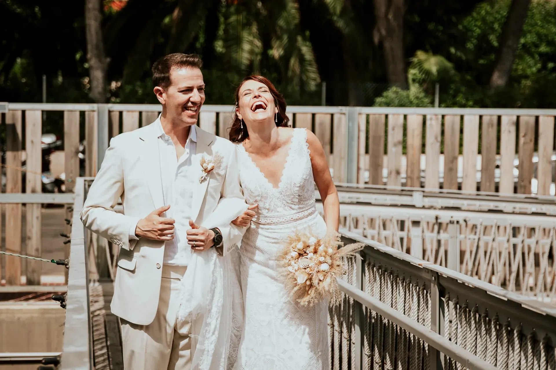 Novios riendo, del brazo, caminando por un puente. El novio lleva un traje color canela, la novia un vestido blanco.