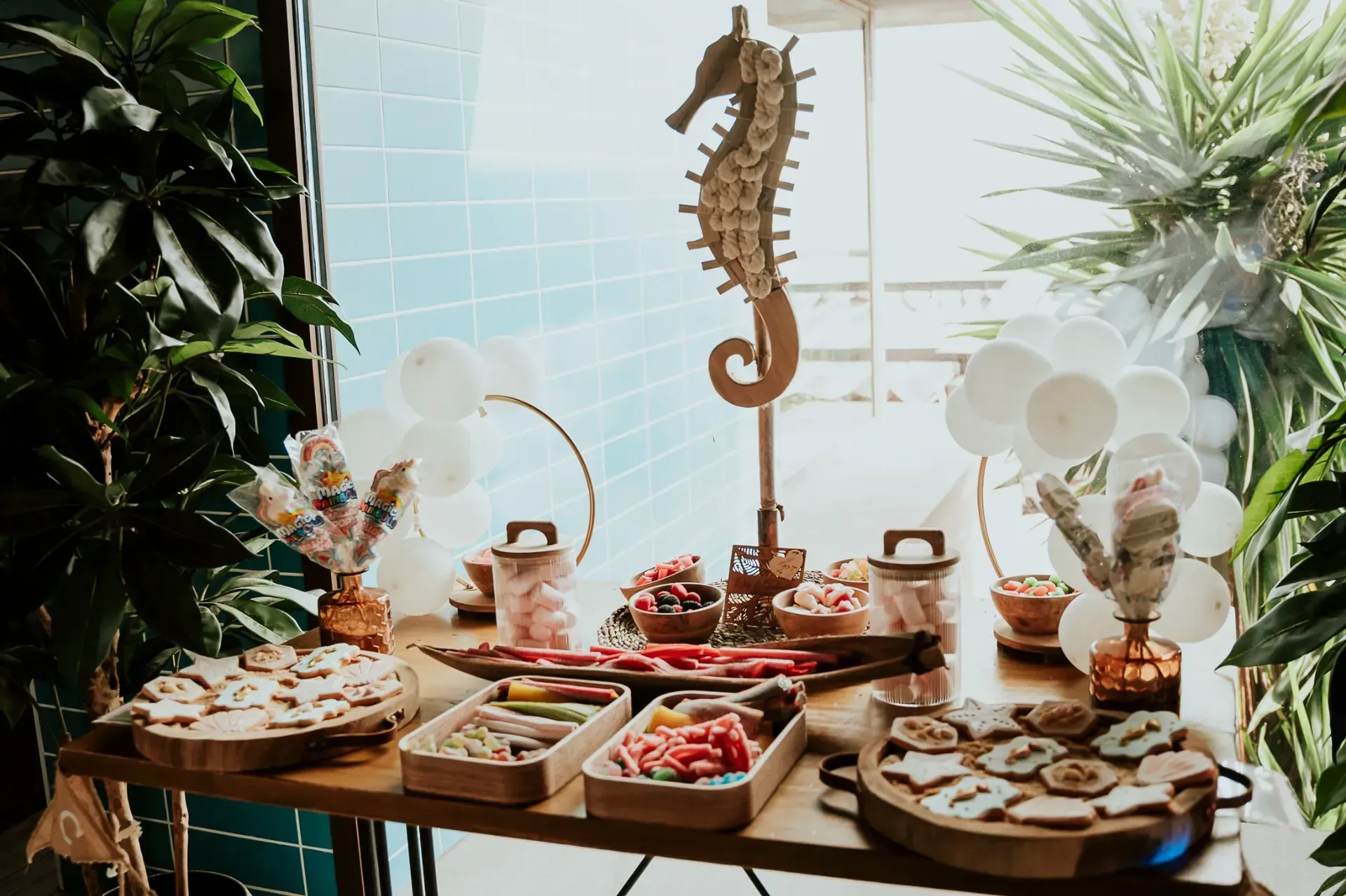 Mesa de buffet de dulces con decoración de caballitos de mar, globos blancos y golosinas en bandejas de madera.