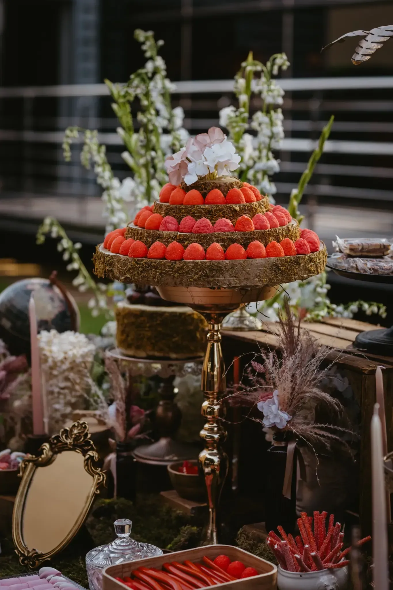Exhibición de postres escalonados con detalles dorados, coronados con dulces rojos, en un entorno de jardín.