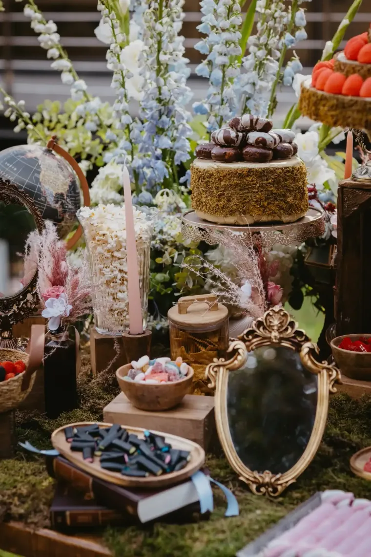 Una mesa de postres con pasteles, dulces, flores, un espejo y un globo terráqueo. Dorado, verde y colores pastel.