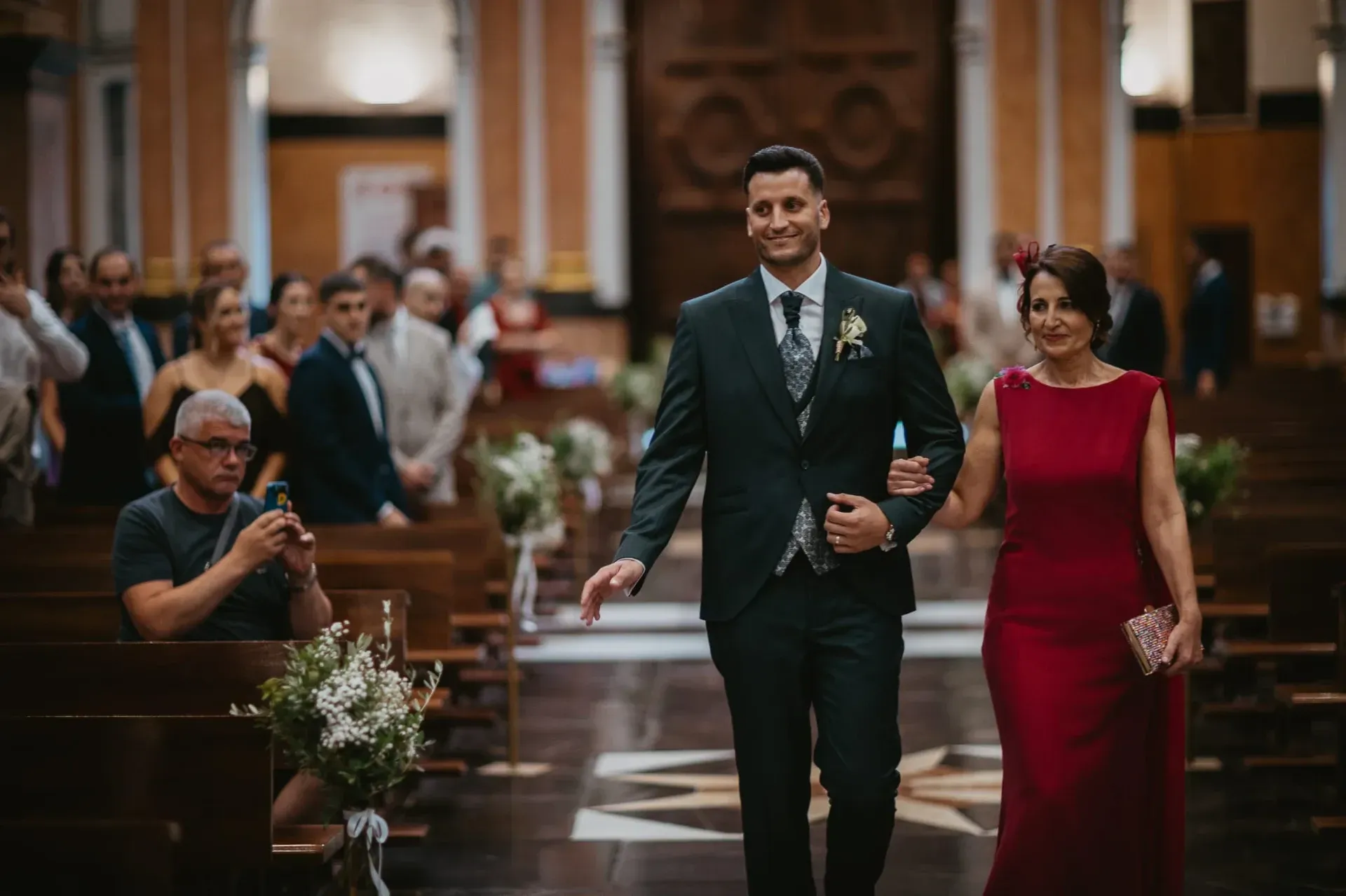 Un hombre con traje camina con una mujer con vestido rojo por el pasillo de la iglesia.