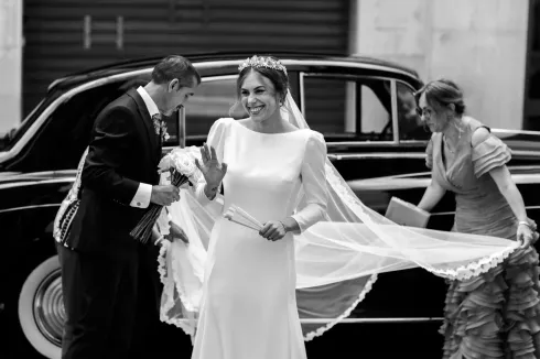 La novia saluda cerca de un coche negro; el novio ajusta la corbata, la mujer ajusta el velo.