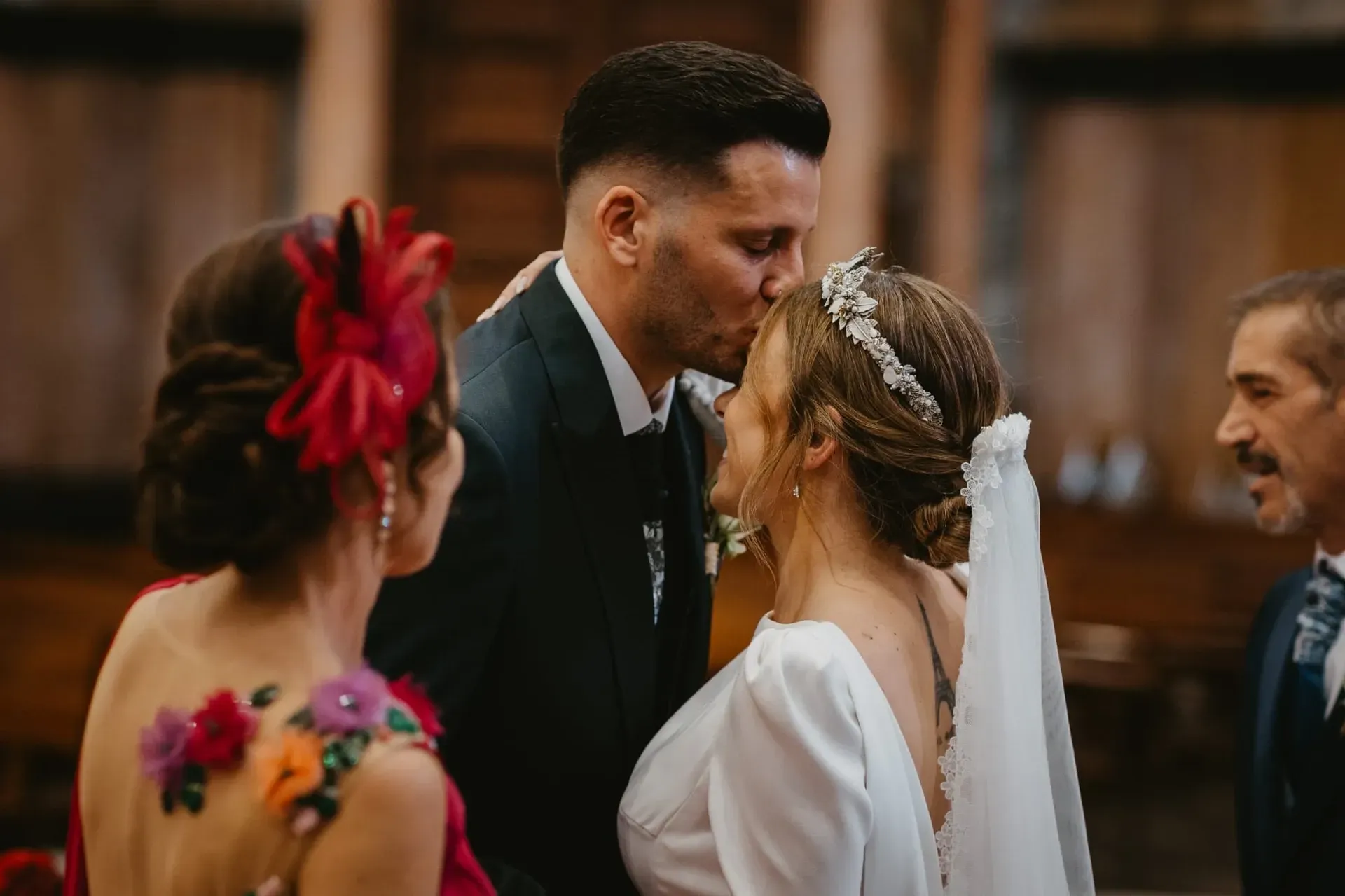 El novio besa la frente de la novia durante una ceremonia nupcial en una iglesia. Los invitados están cerca.
