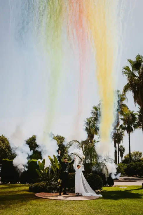 Una pareja bajo una lluvia de polvo de color arcoíris en una ceremonia de boda al aire libre; palmeras al fondo.
