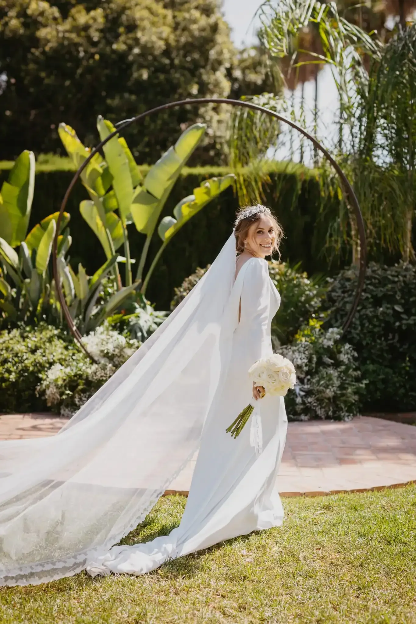 Novia con vestido blanco sosteniendo ramo, sonriendo al aire libre con velo, frente a una estructura circular.