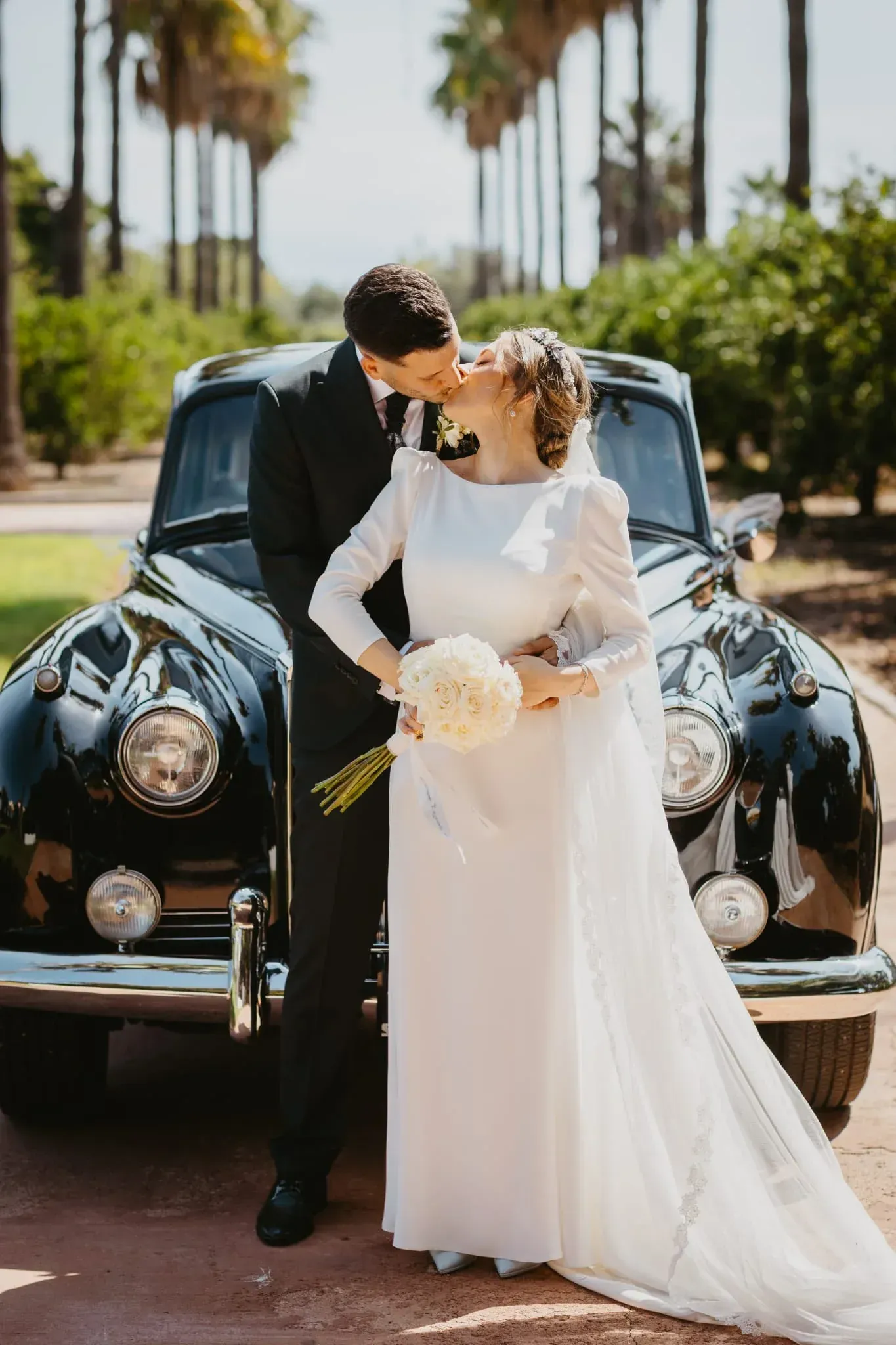 Pareja besándose junto a un coche clásico negro. La novia, con vestido blanco, sostiene un ramo. Fondo con palmeras.